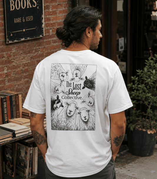 A man viewed from behind wearing a white graphic t-shirt with a black illustration of sheep and the text 'The Lost Sheep Collective' on the back, standing in front of a brick wall and bookshelf.