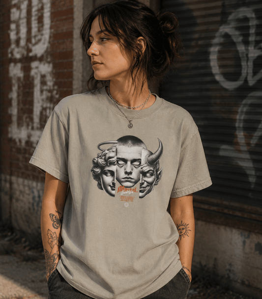 A woman wearing a light gray graphic t-shirt with a black and white illustration of a face with horns and the word 'Morphe' in orange, standing against a graffiti-covered brick wall.