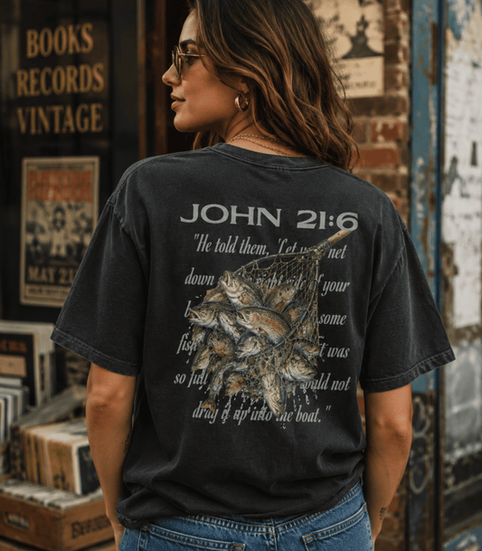 A woman wearing a faded black graphic t-shirt with a fish illustration and the text 'JOHN 21:6' on the back, standing in front of a rustic brick and wood wall.