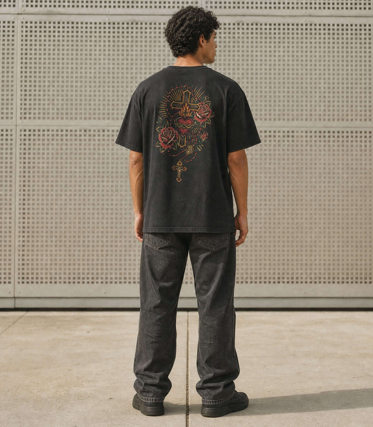 A man viewed from behind wearing a black graphic t-shirt with a large multicolor mandala design on the back, paired with dark gray jeans and black shoes, standing in front of a perforated metal wall.