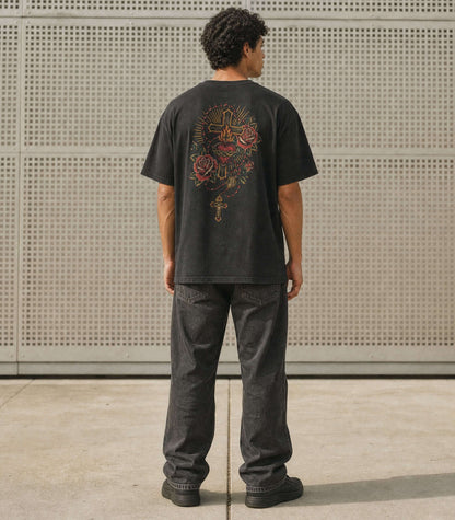 A man viewed from behind wearing a black graphic t-shirt with a large multicolor mandala design on the back, paired with dark gray jeans and black shoes, standing in front of a perforated metal wall.