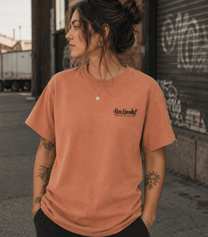 A woman wearing a washed terracotta orange t-shirt with a black 'Hate Loveness' graphic on the left chest, standing in an urban alleyway.