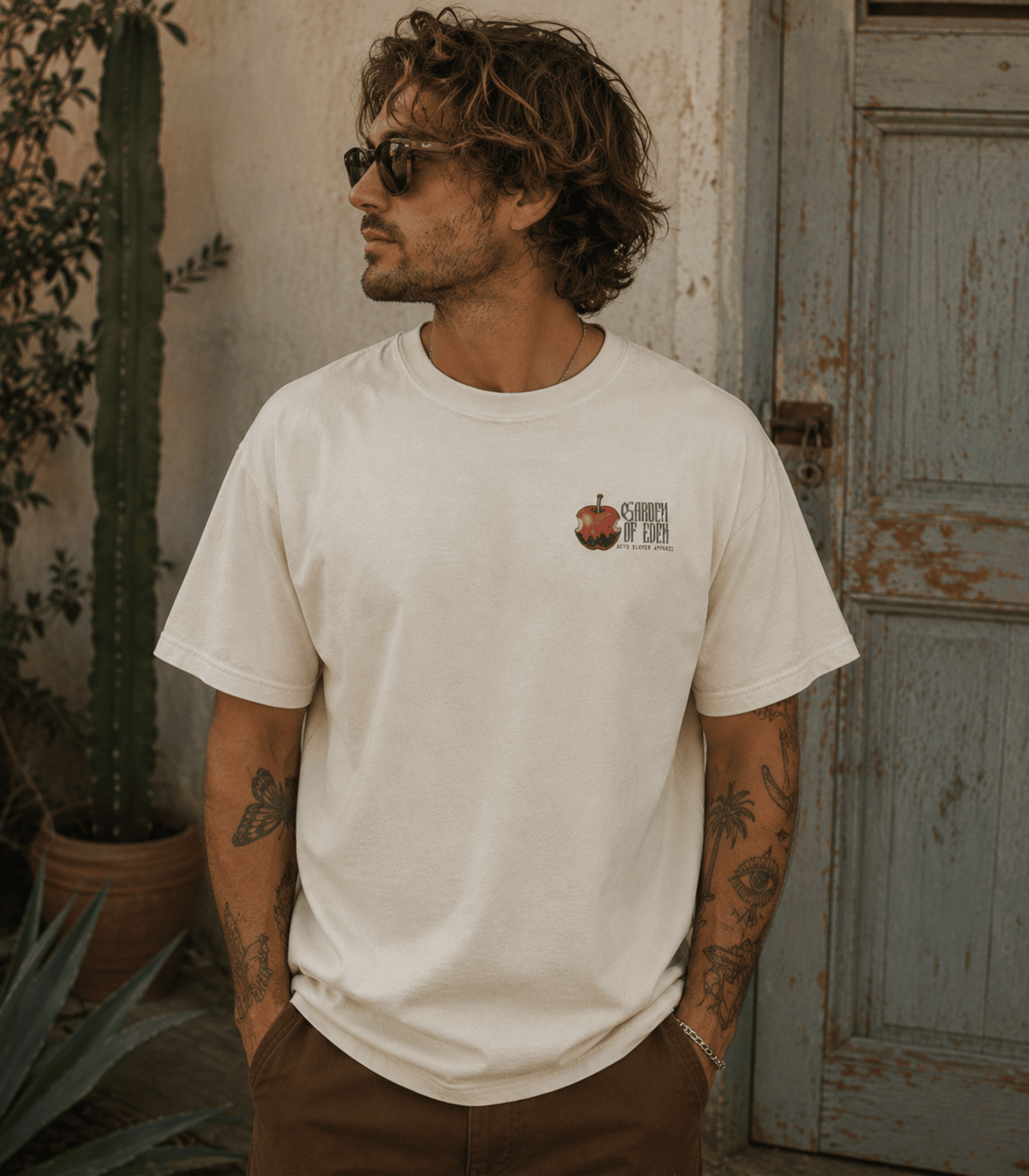 A man wearing a cream-colored crewneck t-shirt with a small graphic on the left chest, standing in front of a rustic weathered door and potted cactus.