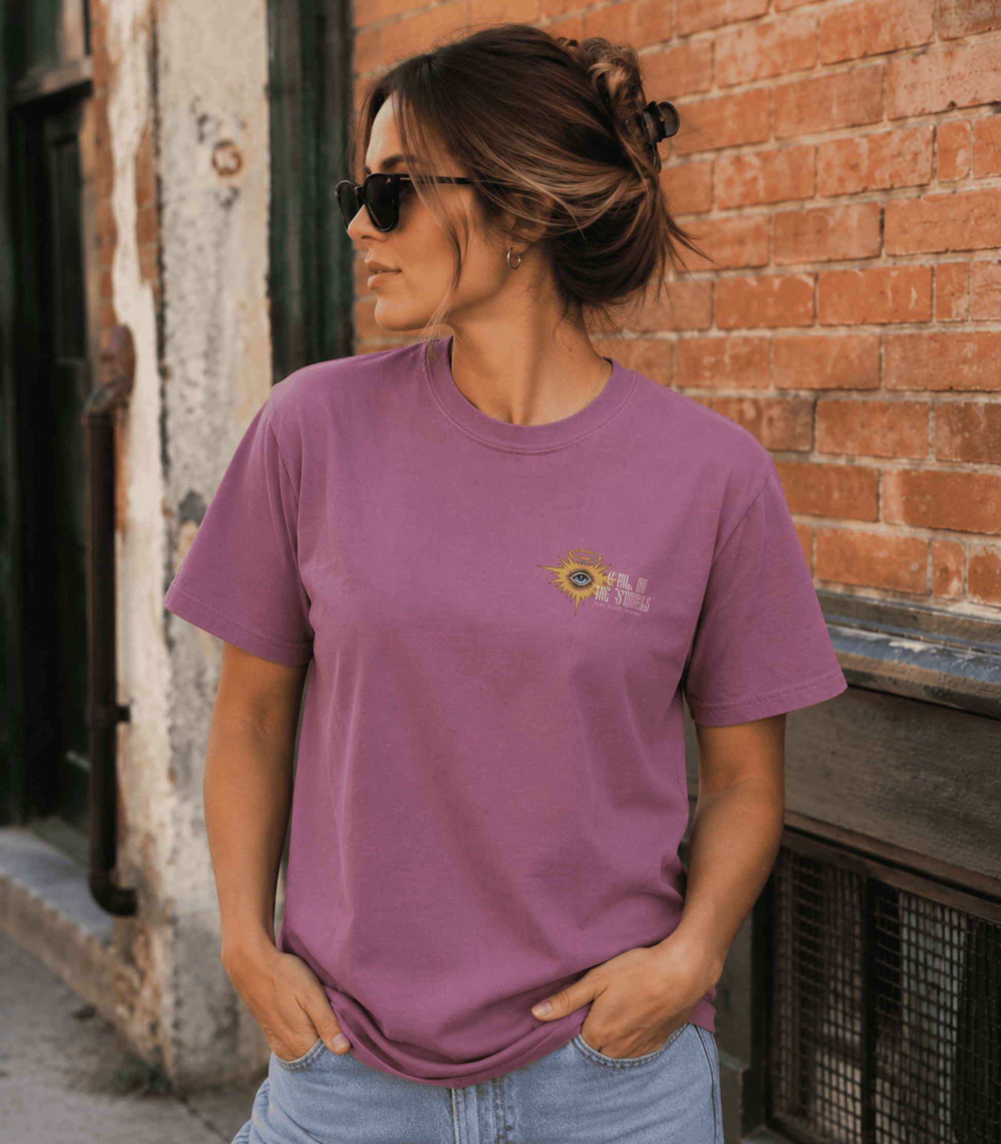 A woman wearing a mauve-purple short-sleeve t-shirt with a small yellow and white graphic on the left chest, paired with light blue jeans, standing against a brick wall.