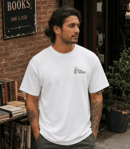 A man wearing a white crewneck t-shirt with a small black graphic on the left chest, standing in front of a brick wall and a bookshop sign.