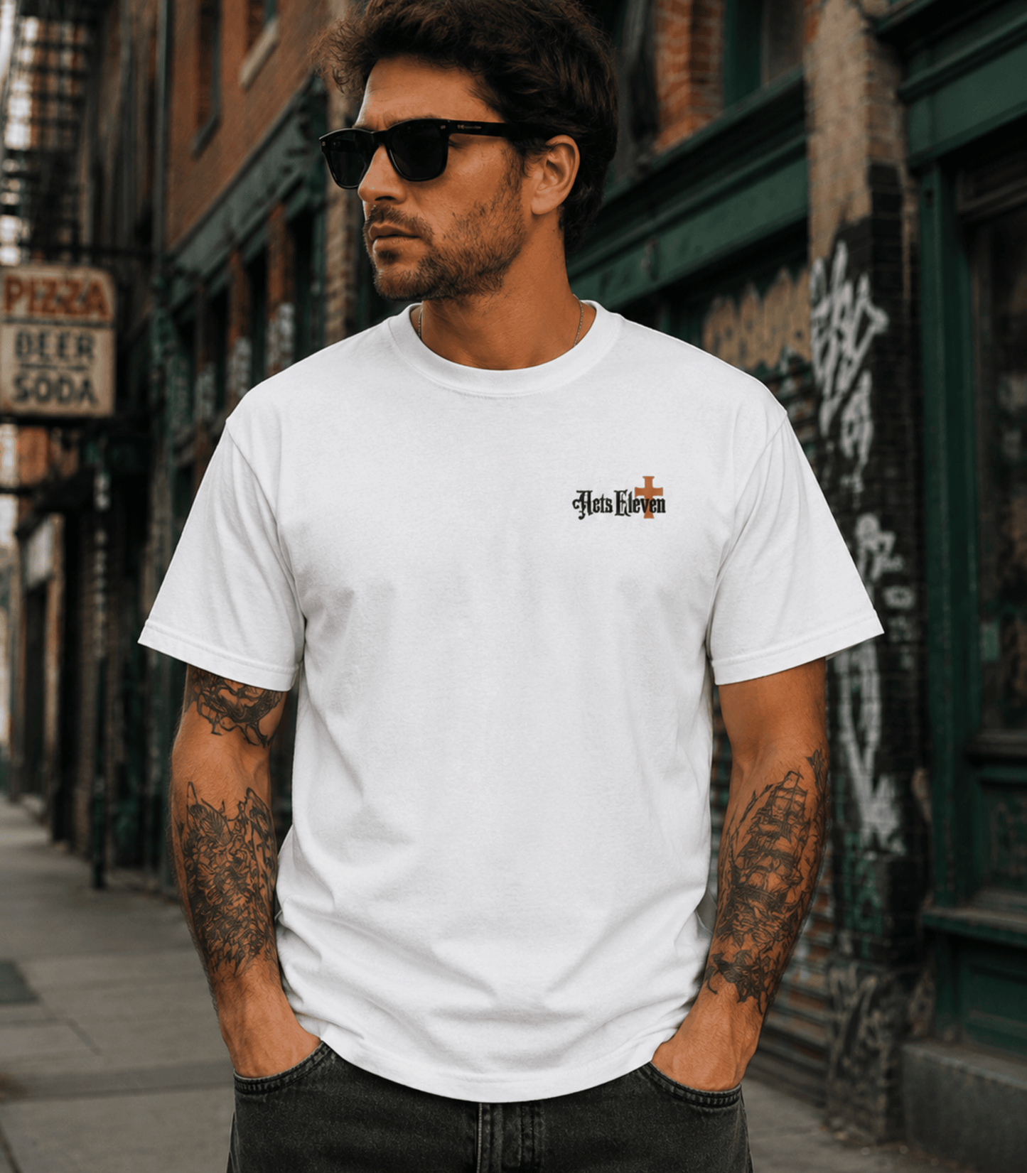 A man wearing a white short-sleeve t-shirt with a small gothic-style 'Heb Eschat' graphic on the left chest, standing on a city street.