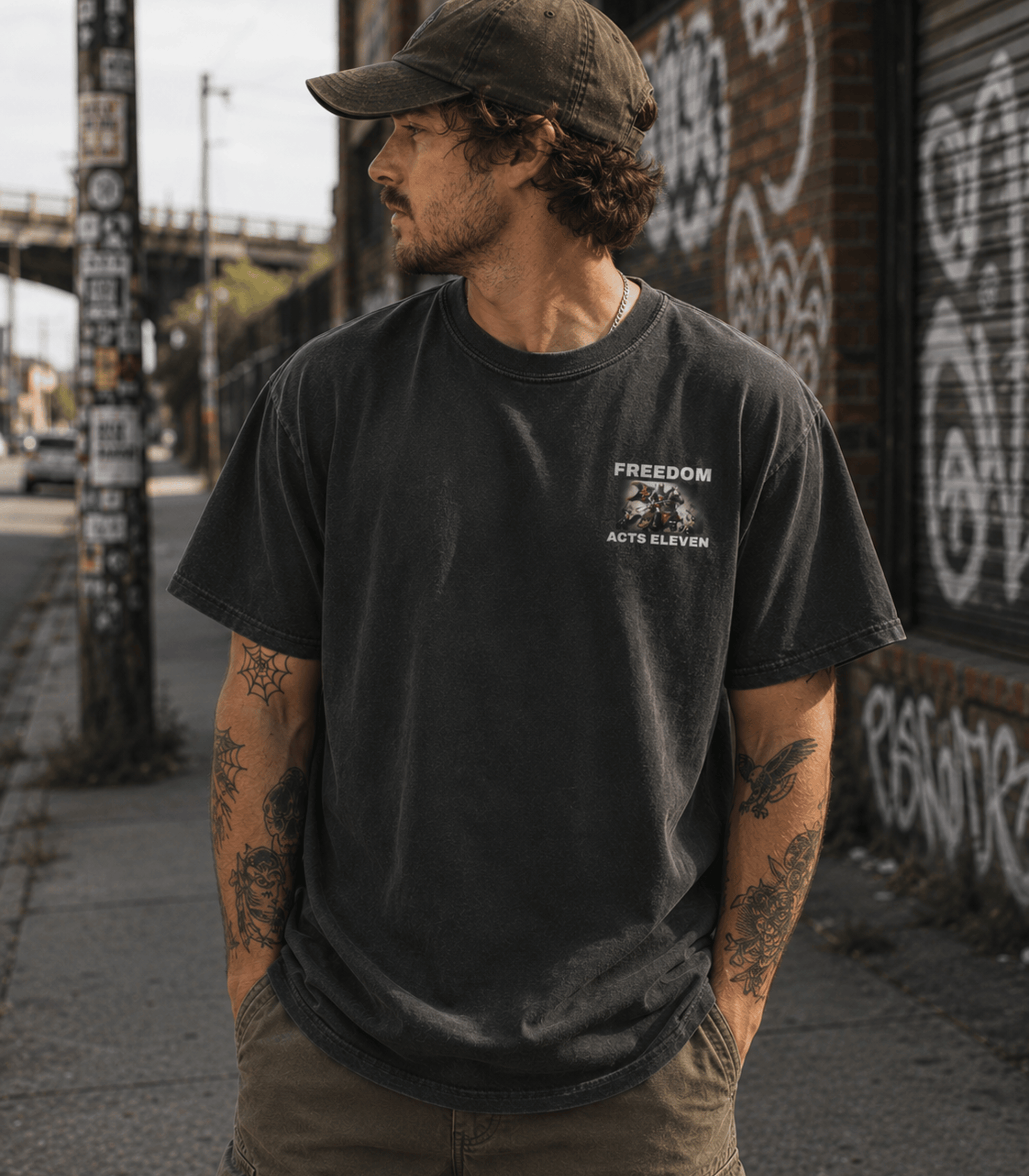 A man wearing a washed black t-shirt with a white 'FREEDOM POLICE ACT 11' graphic on the left chest, paired with a dark cap and khaki pants, standing on an urban sidewalk.
