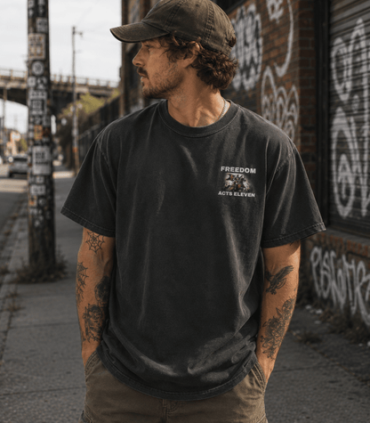 A man wearing a washed black t-shirt with a white 'FREEDOM POLICE ACT 11' graphic on the left chest, paired with a dark cap and khaki pants, standing on an urban sidewalk.