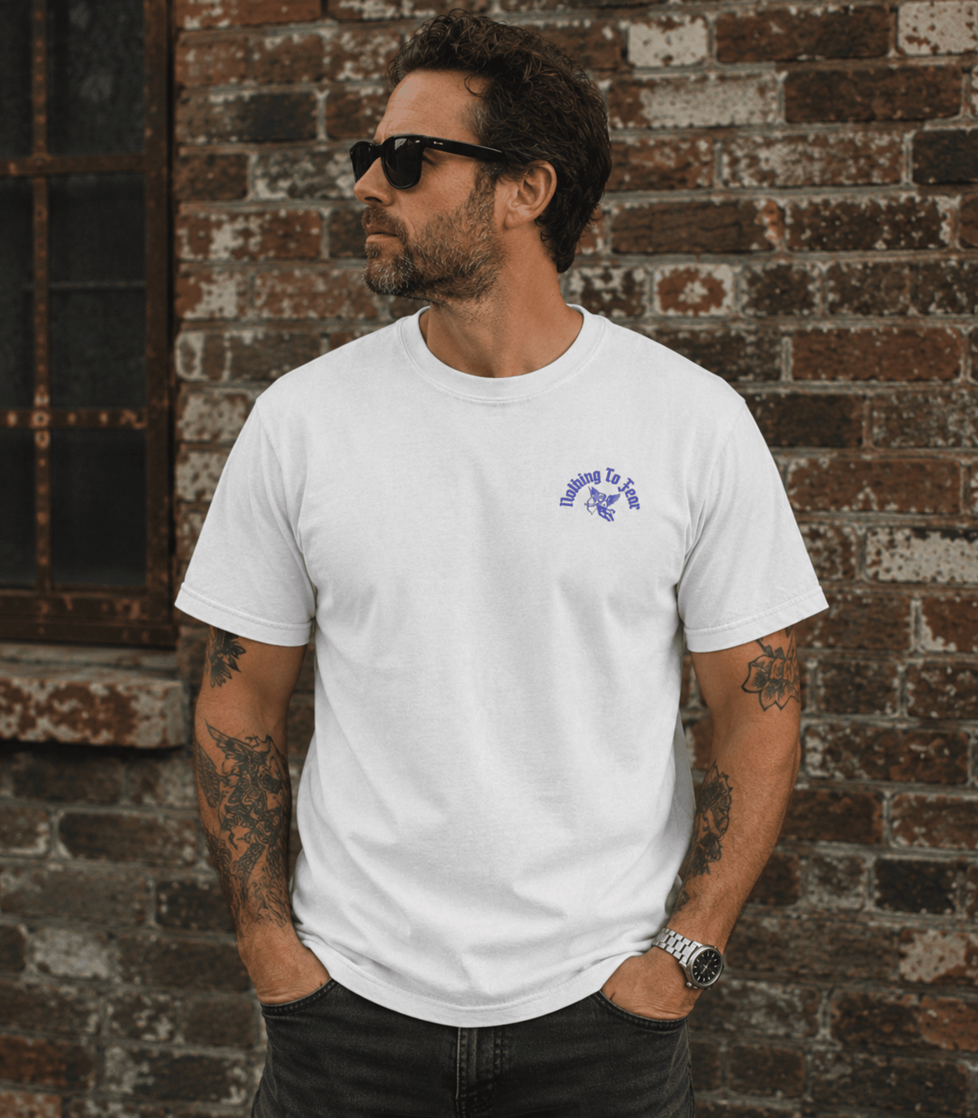 A man wearing a white crewneck t-shirt with a small purple graphic on the left chest, standing in front of a rustic brick wall.