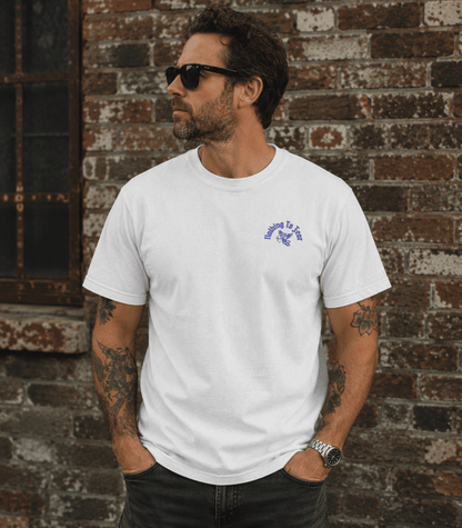 A man wearing a white crewneck t-shirt with a small purple graphic on the left chest, standing in front of a rustic brick wall.