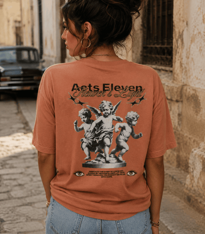 A woman viewed from behind wearing a terracotta-colored graphic t-shirt with 'Acts Eleven' and 'Faith & Loyalty' printed on the back, set against a blurred street background.