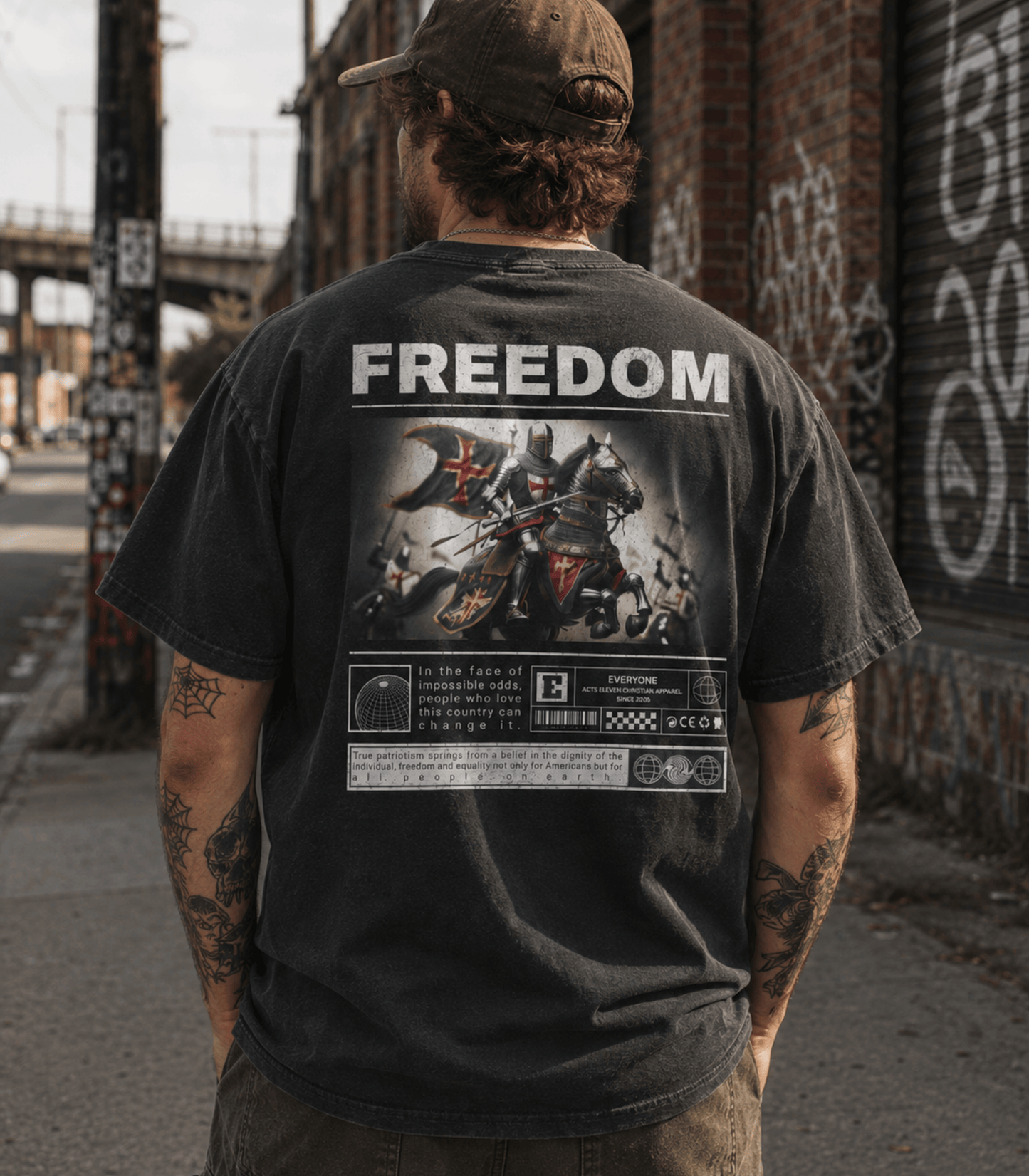A man viewed from behind wearing a faded black graphic t-shirt with a large 'FREEDOM' print and motorcycle illustration on the back, standing on a city street.