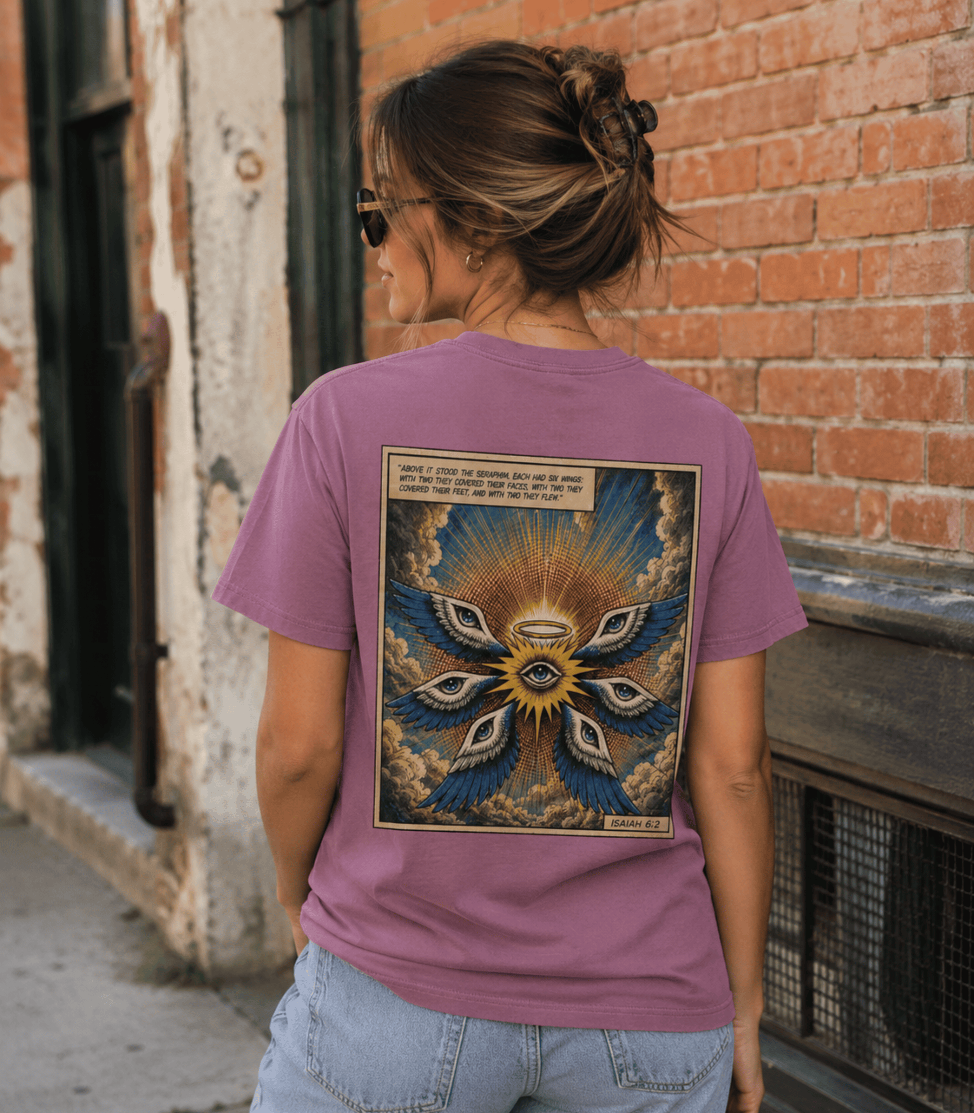 A woman viewed from behind wearing a mauve-purple graphic t-shirt with a large detailed spiritual design on the back, standing outdoors against a brick wall.