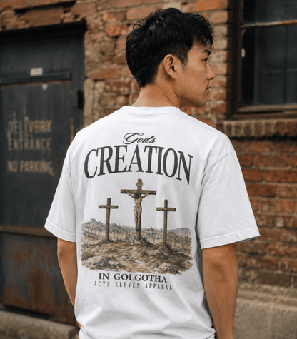 A man wearing a white t-shirt with a large black graphic print on the back, standing in front of a brick wall. The graphic features three crosses and the text 'God's CREATION IN GOLGOTHA' with 'ACCS ELEVEN APPAREL' at the bottom.