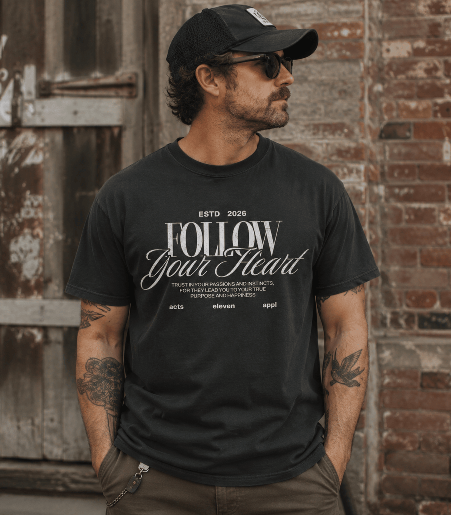 A man wearing a black graphic t-shirt with 'FOLLOW Your Heart' printed in white, paired with a black baseball cap and sunglasses, standing against a rustic brick and wood door background.