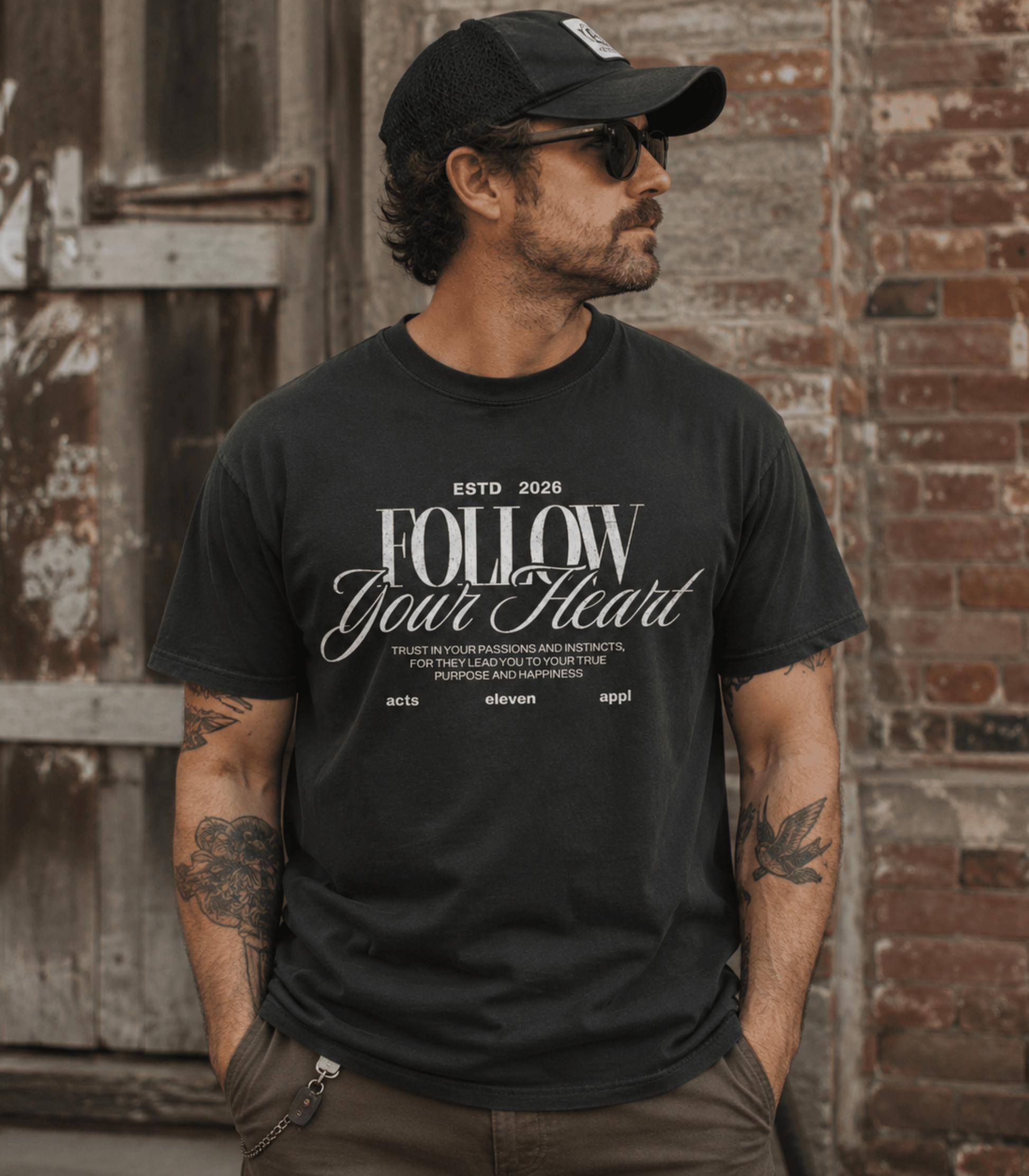 A man wearing a black graphic t-shirt with 'FOLLOW Your Heart' printed in white, paired with a black baseball cap and sunglasses, standing against a rustic brick and wood door background.