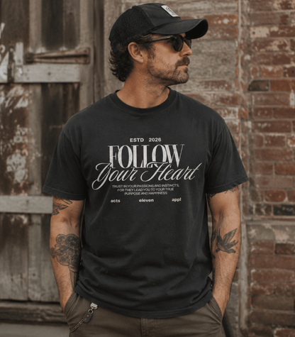 A man wearing a black graphic t-shirt with 'FOLLOW Your Heart' printed in white, paired with a black baseball cap and sunglasses, standing against a rustic brick and wood door background.
