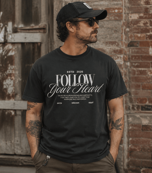 A man wearing a black graphic t-shirt with 'FOLLOW Your Heart' printed in white, paired with a black baseball cap and sunglasses, standing against a rustic brick and wood door background.