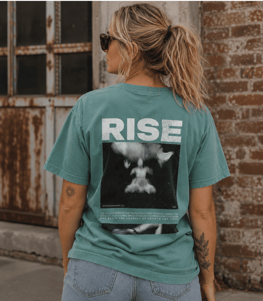 A woman viewed from behind wearing a teal green graphic t-shirt with the band name 'RISE' in white text and a black-and-white photo print on the back, paired with light wash jeans, set against a rustic brick wall and weathered metal door.