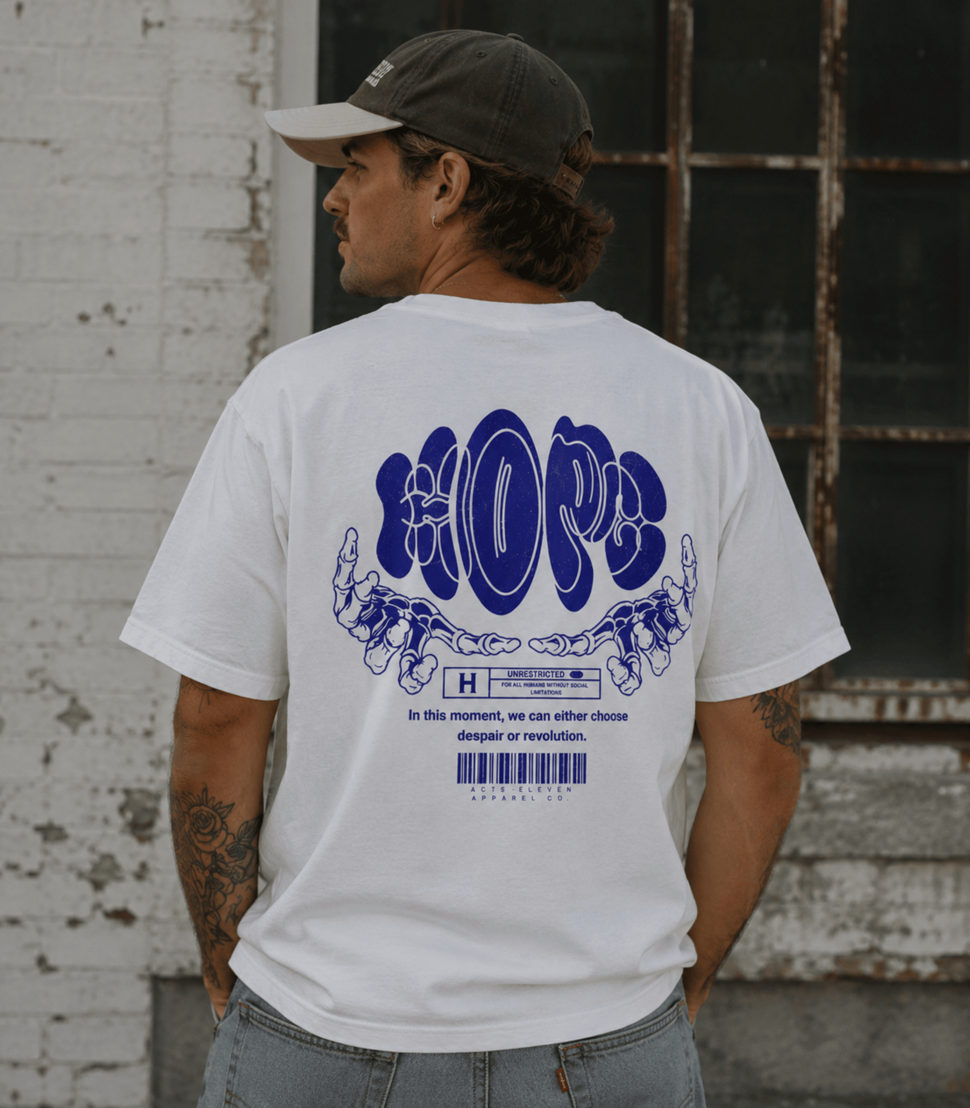 A man wearing a white t-shirt with a large blue graphic on the back, photographed from behind. He is wearing a dark baseball cap and light blue jeans, standing in front of a weathered white brick wall and a rustic window.