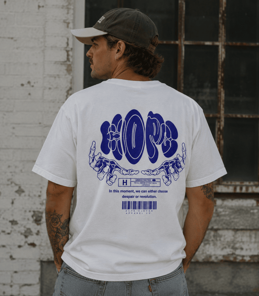 A man wearing a white t-shirt with a large blue graphic on the back, photographed from behind. He is wearing a dark baseball cap and light blue jeans, standing in front of a weathered white brick wall and a rustic window.