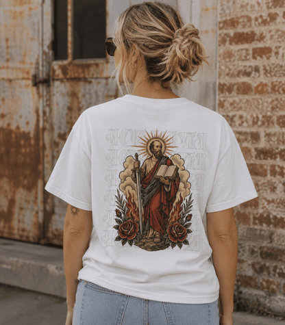 A woman viewed from behind wearing a white graphic t-shirt with a large vintage-style religious illustration of Saint Paul on the back, paired with light blue jeans, set against a rustic brick and metal door background.