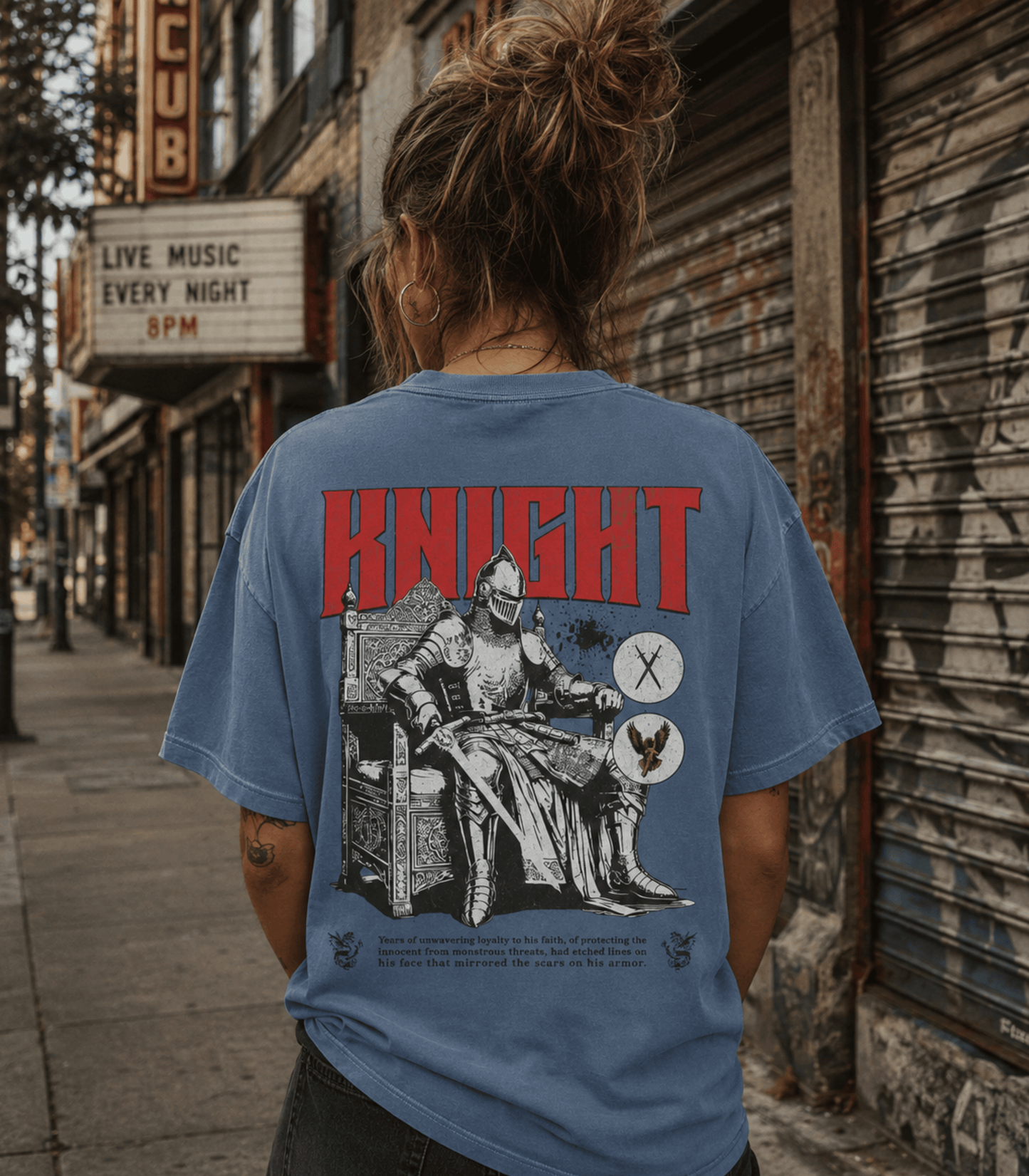A person viewed from behind wearing a faded blue graphic T-shirt on a city sidewalk. The shirt features a large red 'KNIGHT' text and a black-and-white medieval knight illustration with a sword and shield on the back.