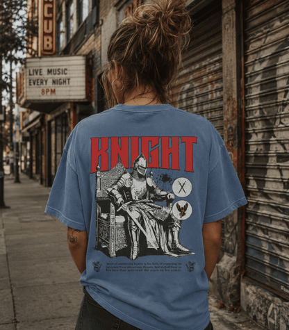 A person viewed from behind wearing a faded blue graphic T-shirt on a city sidewalk. The shirt features a large red 'KNIGHT' text and a black-and-white medieval knight illustration with a sword and shield on the back.