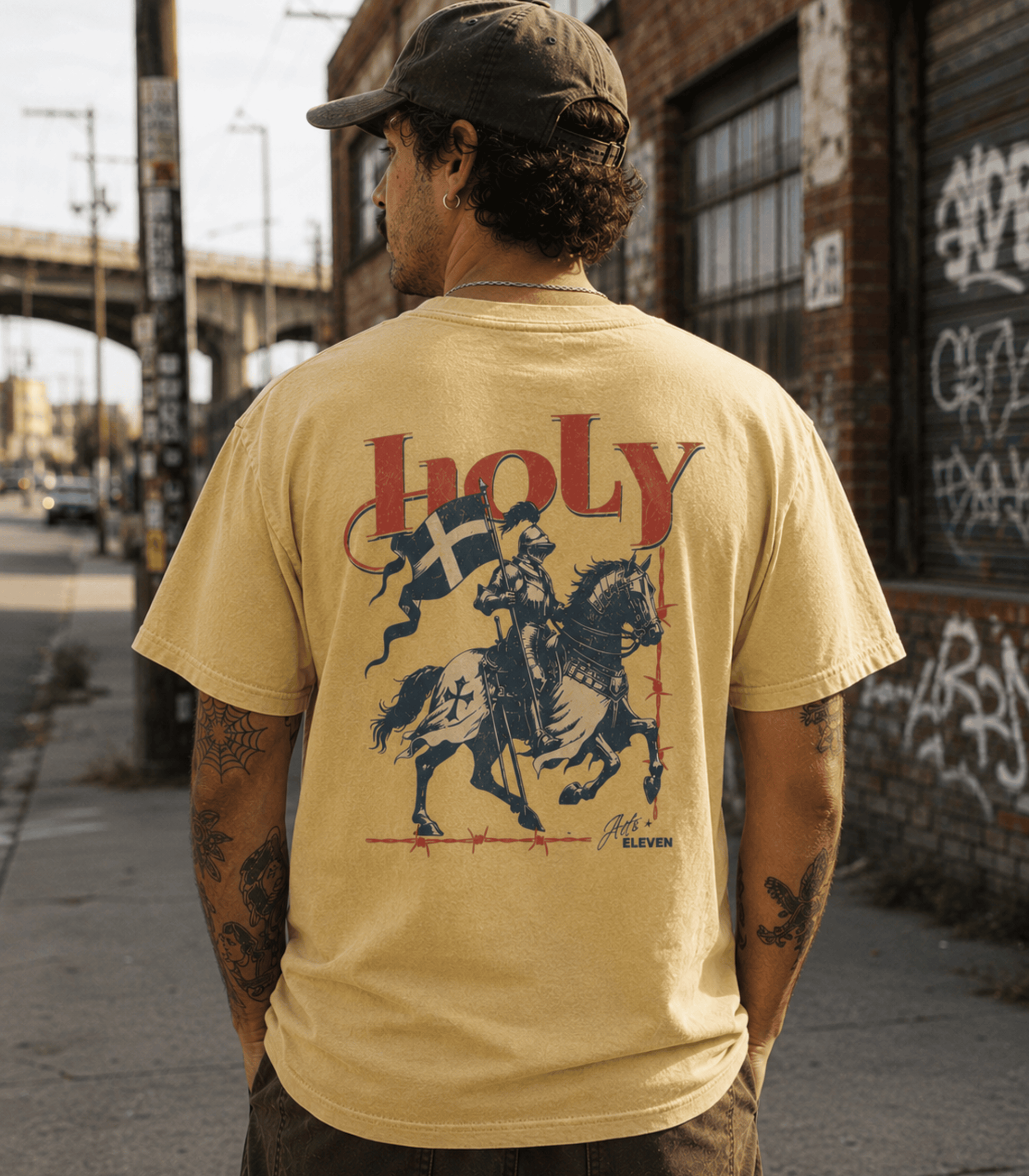A man viewed from behind wearing a tan t-shirt with a large graphic print on the back. The graphic features the word 'holy' in red, a blue and white flag, and a knight on a horse.