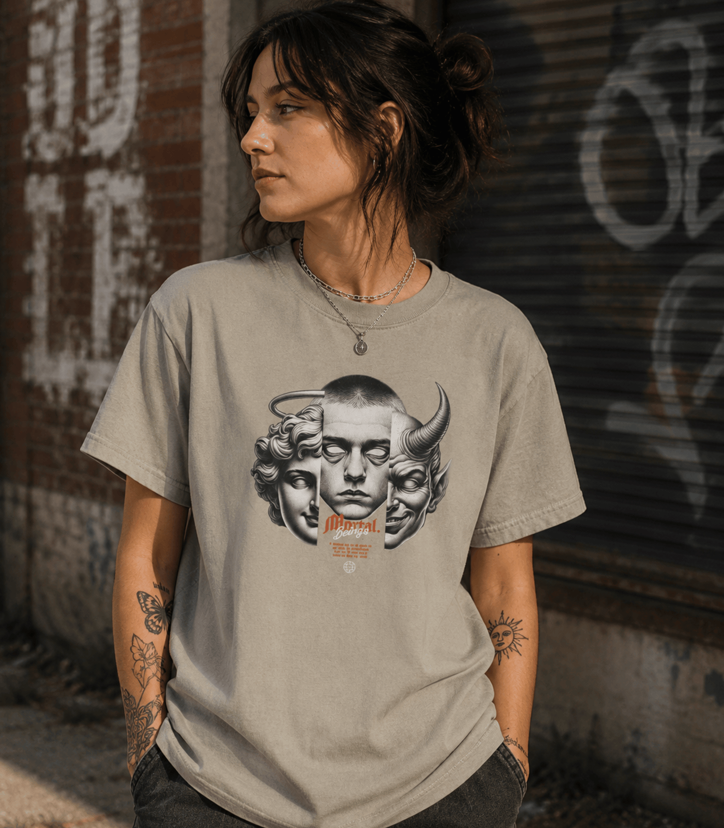 A woman wearing a light gray graphic t-shirt with a black and white illustration of a face with horns and the word 'Morphe' in orange, standing against a graffiti-covered brick wall.