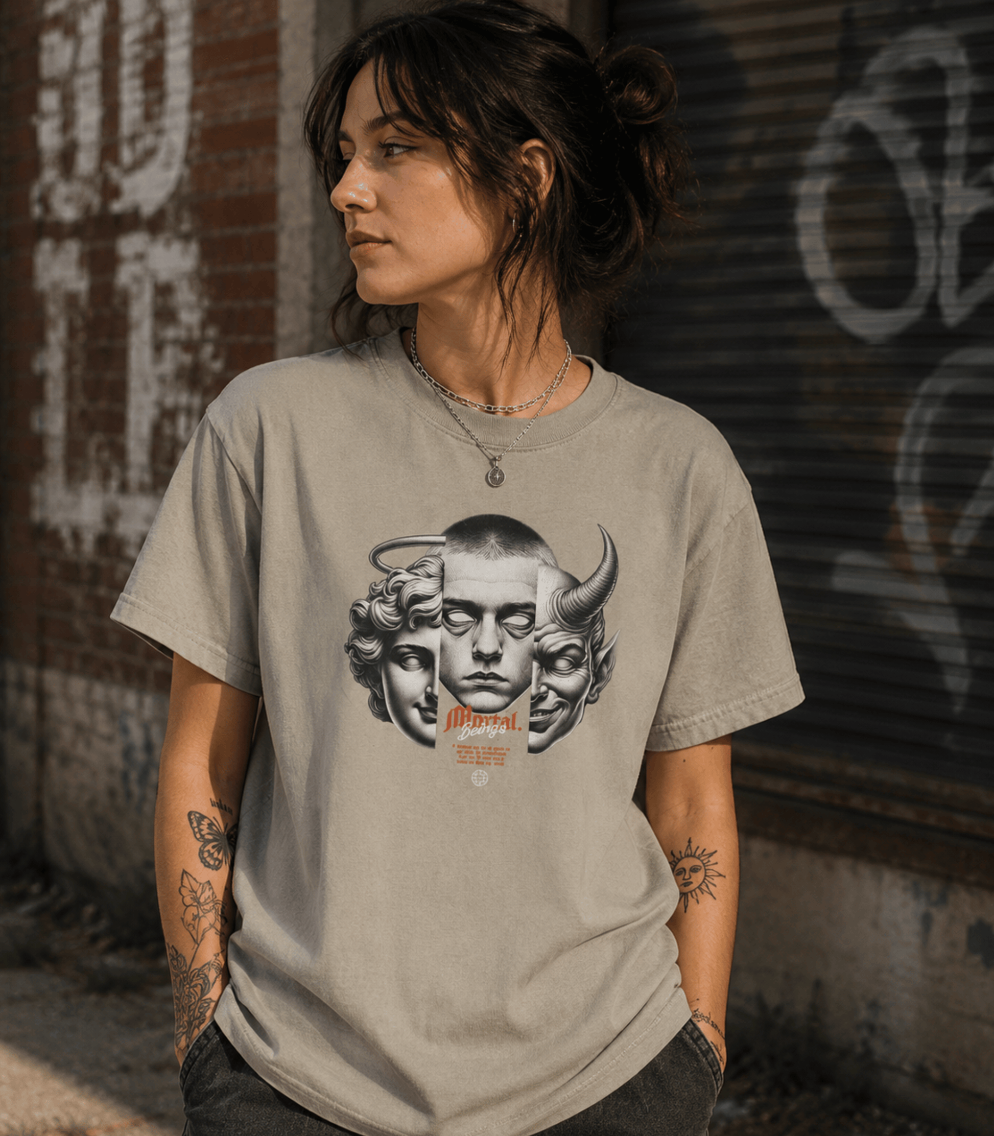 A woman wearing a light gray graphic t-shirt with a black and white illustration of a face with horns and the word 'Morphe' in orange, standing against a graffiti-covered brick wall.