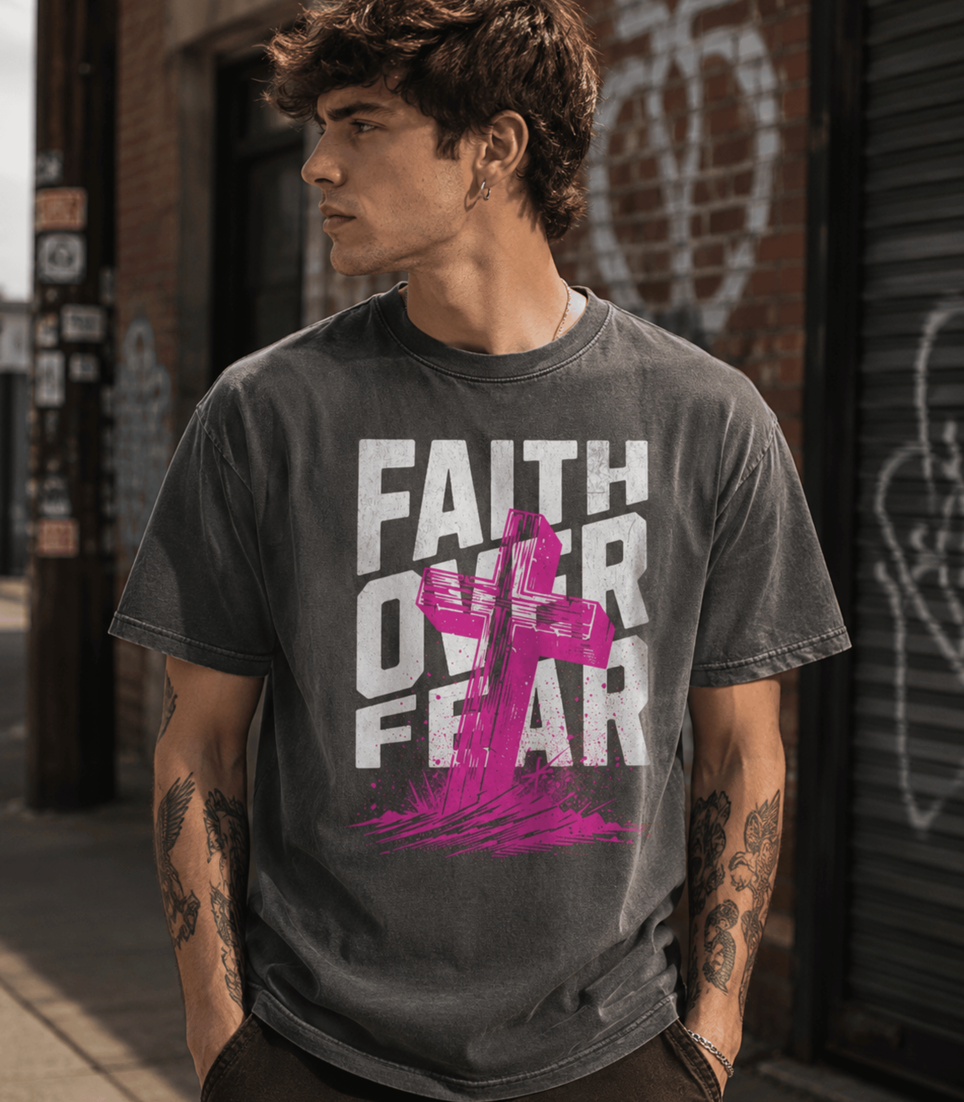 A man wearing a dark gray acid-wash graphic t-shirt with the text 'FAITH OVER FEAR' in white and a pink cross graphic, standing in an urban street setting.