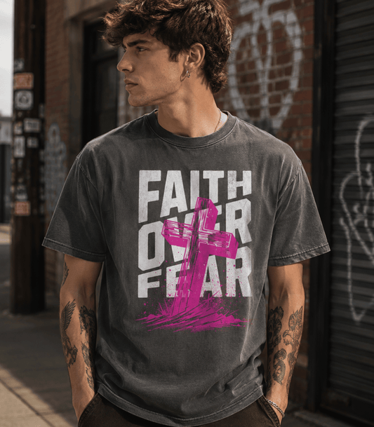 A man wearing a dark gray acid-wash graphic t-shirt with the text 'FAITH OVER FEAR' in white and a pink cross graphic, standing in an urban street setting.