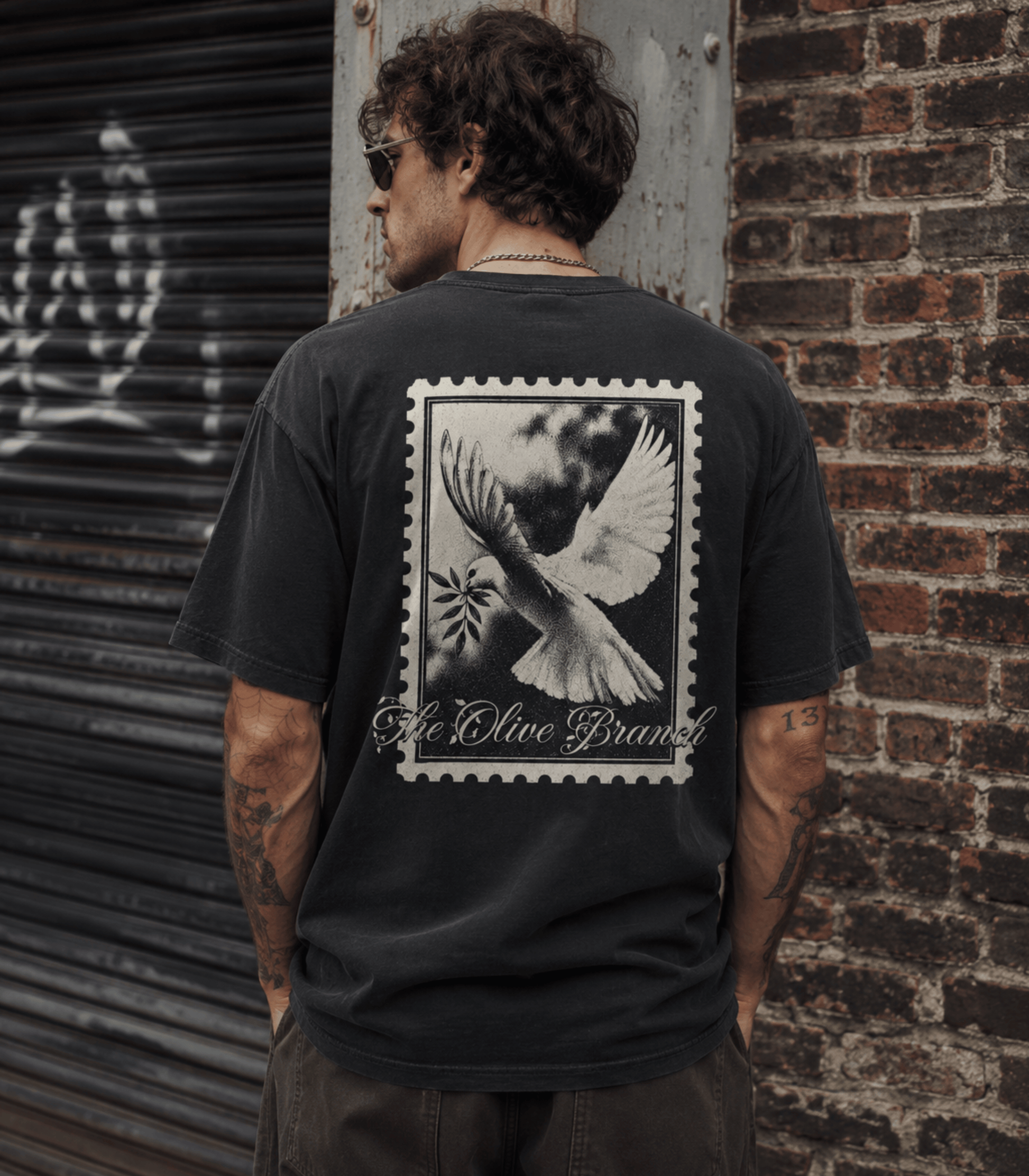 A man wearing a faded black t-shirt with a large white graphic print of a dove and the text 'A Olive Brand' on the back, standing in front of a brick wall and metal shutter.