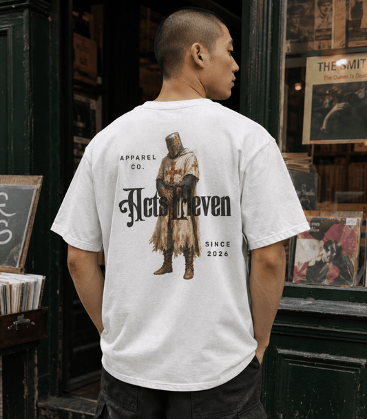 A man viewed from behind wearing a white graphic t-shirt with a large medieval knight design and the text 'Acts Reven' and 'Apparel Co.' printed on the back, standing in front of a dark shop window.