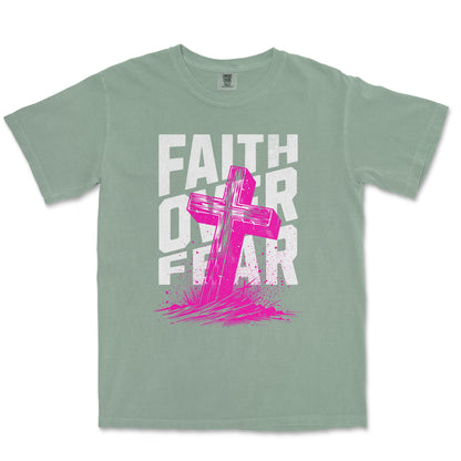 A sage green short-sleeve t-shirt with a graphic print on the front featuring the words 'FAITH OVER FEAR' in distressed white letters and a bright pink cross layered over them.